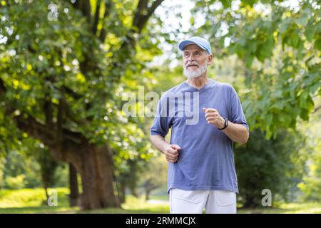 Homme âgé aux cheveux gris jogging dans le parc, retraité souriant contentement reposant dans le parc parmi les arbres, portant des vêtements de sport et une casquette sur sa tête. Banque D'Images