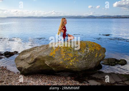 Une belle femme blonde, Rhianna Martin, est assise sur les rochers de la plage de Wormit dans le comté de Fife, en Écosse, profitant du temps agréable, au Royaume-Uni Banque D'Images