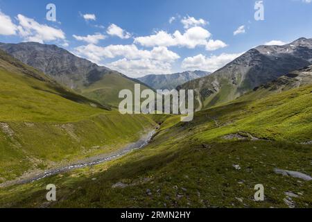 Un ruisseau de montagne traverse une large et verte haute vallée sur le chemin des prairies de Kvakhidi au col d'Atsunta dans la municipalité d'Akhmeta, région de Kakhétie, Géorgie. Des pentes herbeuses s'élèvent des deux côtés. À droite, un sentier étroit monte en diagonale sur la pente. En arrière-plan, la vue s'ouvre sur une vallée latérale avec des pics rocheux gris. Les prairies se situent à une altitude d'environ 2 400 mètres dans la haute vallée de Kvakhidistskali du parc national de Tusheti. Le sentier de Girevi au col d'Atsunta traverse cette vallée et le village abandonné de Chontio. Les Tushetiens semi-nomades vivent dans Banque D'Images