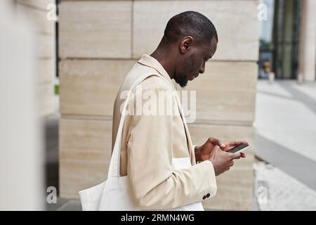 Manager masculin afro-américain en blanc formalwear texting dans smartphone tout en se tenant devant la caméra par un bâtiment moderne dans la rue Banque D'Images