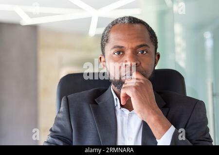 Homme afro-américain mûr focalisé regardant la caméra, gros plan portrait de patron concentré réfléchi au lieu de travail à l'intérieur du bureau en costume d'affaires. Banque D'Images