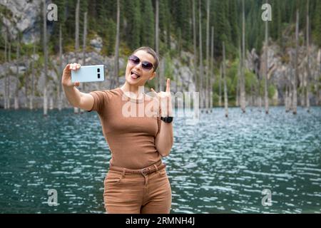 Une jeune femme voyage à travers les montagnes de Tien Shan en Asie centrale, fait un selfie joyeux sur fond de lac de montagne pittoresque Banque D'Images