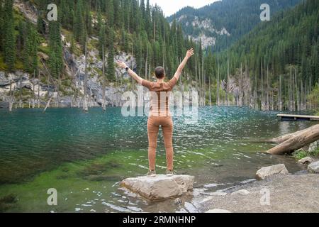 Femme touristique voyage à travers les montagnes de l'Asie centrale, debout avec les bras tendus et regardant le lac de montagne Kaindy avec le sapin coulé Banque D'Images