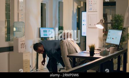 Comptable afro-américain se faufilant sur un collègue endormi sieste à son bureau pendant le quart de nuit. Employé de bureau drôle jouant à la blague sur le collègue endormi, ajustant son niveau de hauteur de chaise par derrière Banque D'Images