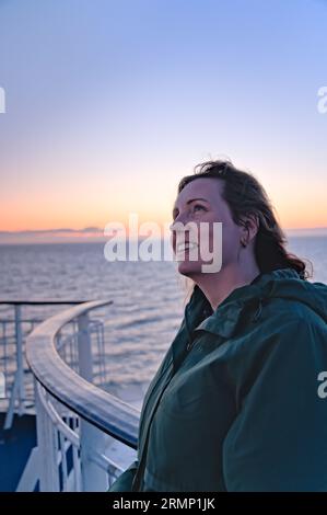 Belle femme appuyée contre la balustrade de bateau regardant le ciel lumineux de coucher de soleil avec un sourire sur son visage et avec le vent dans ses cheveux.mer scandinave Banque D'Images