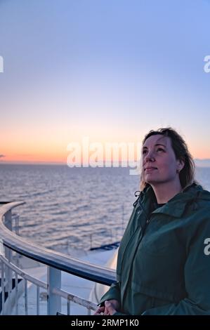 Beautiful woman leaning against the railing of the boat, looking at the sky with a satisfied face with the wind in her hair. Glowing sunset on the sea Stock Photo