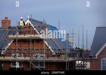 Nouvelles propriétés résidentielles à différents stades de construction : maisons avec des toits complètement finis, une partiellement carrelée, et des hommes travaillant sur des échafaudages. Banque D'Images