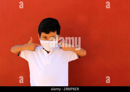 Joyeux et pensif Latino garçon de 8 ans portant une chemise blanche d'uniforme scolaire et un masque facial pour la protection contre la pandémie de Covid-19 à la rentrée scolaire Banque D'Images