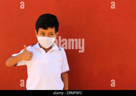 Joyeux et pensif Latino garçon de 8 ans portant une chemise blanche d'uniforme scolaire et un masque facial pour la protection contre la pandémie de Covid-19 à la rentrée scolaire Banque D'Images