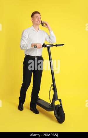 Homme heureux avec scooter de kick électrique moderne parlant sur smartphone sur fond jaune Banque D'Images