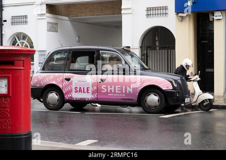 Un taxi noir londonien traditionnel avec une publicité, à côté d'un coureur de scooter de livraison de nourriture, avec un cycliste derrière et un pilarbox rouge. Londres, Royaume-Uni Banque D'Images