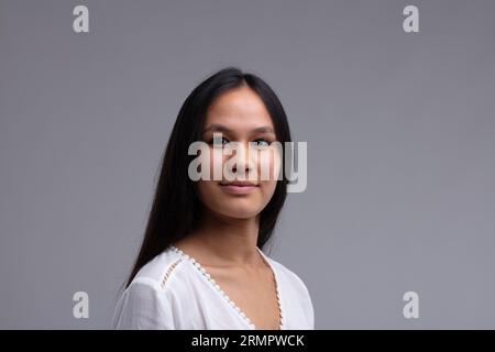 Portrait rapproché de souriant belle femme brune avec des cheveux noirs épais, portant un chemisier tricoté blanc. Portrait de buste demi-tour sur fond gris Banque D'Images