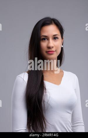 Jolie femme aux cheveux foncés portant des boucles d'oreilles en perle et un haut en dentelle blanche avec ses cheveux balayés sur une épaule regardant la caméra sur gris Banque D'Images