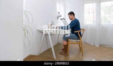 Un homme avec une jambe cassée immergé dans son travail, assis dans un bureau ou un poste de travail à domicile. Il est absorbé par diverses tâches sur son ordinateur portable, le transport Banque D'Images