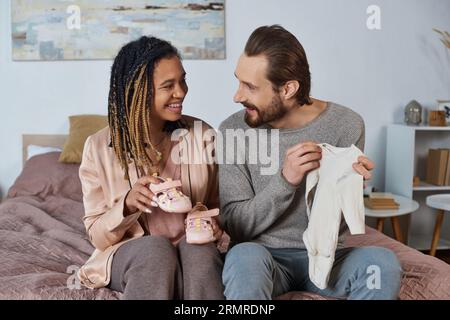 heureux couple interracial tenant des vêtements de bébé mignons, chaussures minuscules, futurs parents, attente Banque D'Images