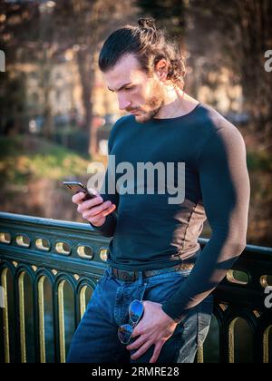 Homme musclé avec les cheveux longs en tenue décontractée regardant l'écran de mobile et de messagerie texte tout en se relaxant dans le parc vert Banque D'Images
