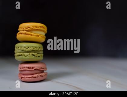 Macarons de couleurs jaune, rose et vert empilés sur marbre blanc et fond noir. Banque D'Images