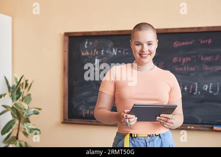 Heureuse jeune enseignante ou étudiante du lycée en t-shirt rose à l'aide de tablette tout en se tenant contre le tableau noir et en regardant la caméra Banque D'Images