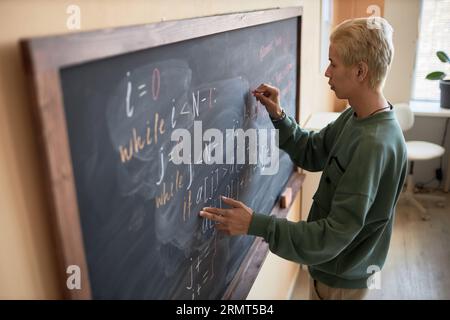 Vue de côté d'un jeune homme blond en casualwear écrivant des formules ou des équations au tableau noir avec de la craie tout en expliquant quelque chose en leçon Banque D'Images