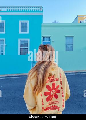 Une jeune femme blonde portant un sweat à capuche jaune se tient devant un bâtiment bleu dans le quartier culturellement dynamique de Bo-Kaap Banque D'Images