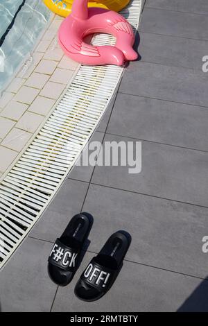 Sandales de piscine avec un message grossier distinctif laissé à côté d'une piscine dans une station de vacances européenne, Simmer 2023 Banque D'Images