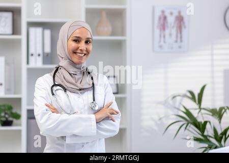 Portrait de jeune femme médecin portant le hijab, femme musulmane souriant et regardant la caméra avec les bras croisés en manteau médical blanc avec stéthoscope, travaillant à l'intérieur du bureau médical.. Banque D'Images