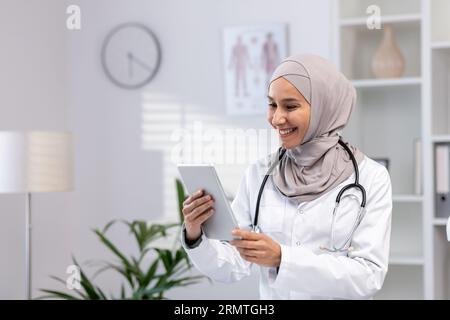 Jeune belle femme médecin dans le hijab à l'aide d'un ordinateur tablette, médecin travailleur en manteau médical blanc et stéthoscope à l'intérieur du bureau de la clinique au travail, souriant femme musulmane à l'aide de l'application en ligne. Banque D'Images