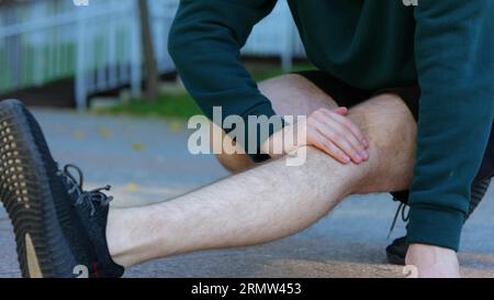 Sportif assis et étirant son genou, jambe, muscles dans le parc vert. Jeune homme caucasien souffrant de fortes douleurs à la jambe. Visage de gars montrant Banque D'Images