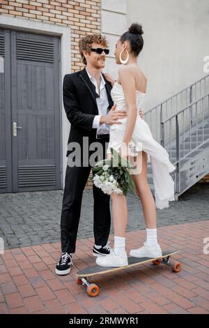 redhead groom dans des lunettes de soleil serrant jeune mariée afro-américaine avec des fleurs debout sur longboard Banque D'Images