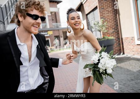 mariée afro-américaine avec des fleurs et un marié roux dans des lunettes de soleil tenant les mains sur la rue Banque D'Images