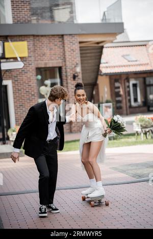 jeune mariée afro-américaine tenant la main avec le marié et chevauchant longboard sur la rue, bonheur Banque D'Images