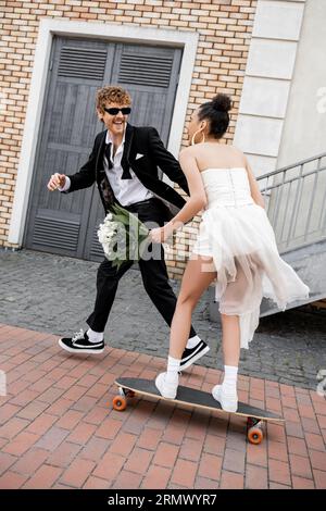 marié excité dans des lunettes de soleil près de mariée afro-américaine en robe de mariée chevauchant longboard Banque D'Images