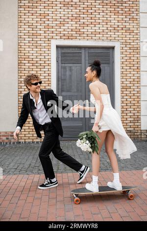 redhead groom dans les lunettes de soleil tenant la main avec la mariée afro-américaine avec bouquet riding longboard Banque D'Images