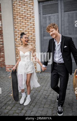 joyeux jeunes mariés interraciaux marchant avec longboard et skateboard dans la rue de la ville, tenue de mariage Banque D'Images