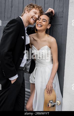 redhead groom et élégante mariée afro-américaine avec skateboard riant près du bâtiment dans la ville Banque D'Images