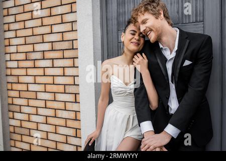 mariée afro-américaine ravie embrassant heureux marié rousse près du bâtiment de la ville, les yeux fermés Banque D'Images