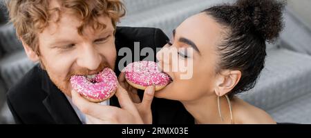 élégant couple interracial se nourrissant les uns les autres avec des beignets doux, mariage dans la rue, plaisir, bannière Banque D'Images