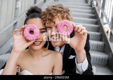 s'amuser, se marier en ville, excités jeunes mariés interraciaux obscurcissant le visage avec des beignets Banque D'Images