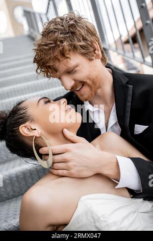 romance urbaine, mariage, tendresse, heureux marié roux embrassant élégante mariée afro-américaine Banque D'Images