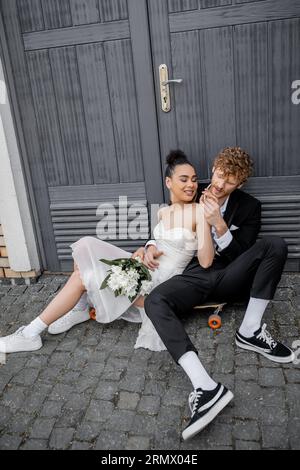 homme de redhead tenant les mains de la mariée afro-américaine avec des fleurs, assis sur le trottoir près des portes Banque D'Images