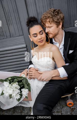le marié redhead embrasse la mariée afro-américaine avec les yeux fermés et les fleurs, assis près du bâtiment Banque D'Images