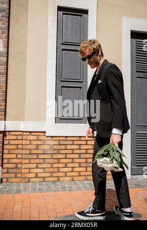 homme élégant en costume noir et lunettes de soleil marchant dans la rue de la ville, marié avec bouquet de mariage Banque D'Images