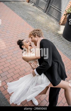 jeune marié élégant serrant mariée afro-américaine excitée, mariage dans la ville européenne Banque D'Images