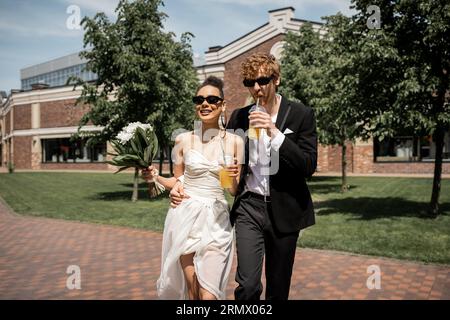 élégants jeunes mariés dans des lunettes de soleil marchant avec du jus d'orange et des fleurs dans la ville européenne Banque D'Images