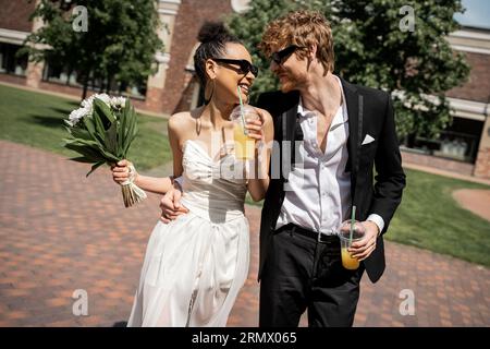 joyeux jeunes mariés multiethniques dans des lunettes de soleil marchant avec du jus d'orange, mariage dans la rue urbaine Banque D'Images