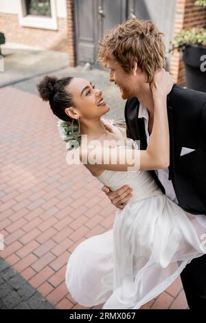 overjoyed et élégant couple interracial en tenue de mariage embrassant sur la rue urbaine Banque D'Images