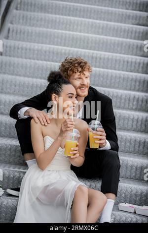 redhead groom embrassant heureuse mariée afro-américaine assise avec du jus d'orange sur les escaliers dans la ville Banque D'Images