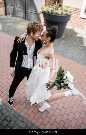amour en ville, marié rousse et femme afro-américaine avec bouquet embrassant sur la rue Banque D'Images