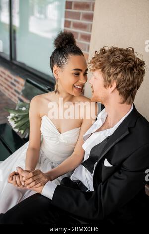 joyeux couple interracial se tenant la main et se regardant sur le banc, mariage en ville Banque D'Images