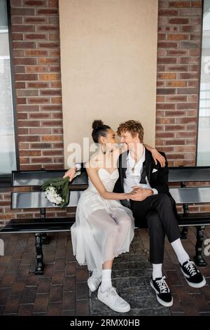 mariage dans la ville, heureux marié roux avec bouquet assis face à face avec la mariée afro-américaine Banque D'Images